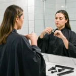 Femme se coupant les cheveux seule dans une salle de bain moderne avec des ciseaux professionnels et une cape de coiffure