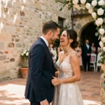Couple de mariés riant sous un arc de pierre fleuri dans une cour provençale lors de leur cérémonie en plein air