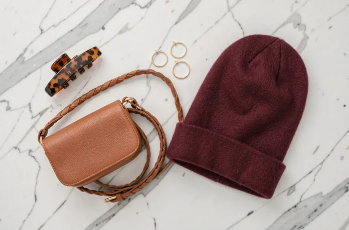 Plat posé d'accessoires de mode tendance sur marbre blanc : clip tortoise, boucles d'oreilles dorées, ceinture tressée, sac cognac et bonnet bordeaux